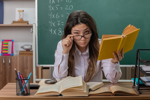 Professora jovem usando óculos com livro concentrado, sentada na mesa da escola em frente ao quadro-negro na sala de aula