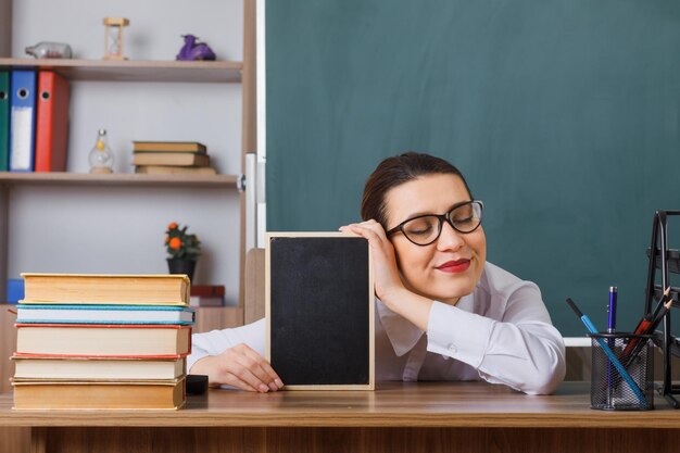 Professora jovem usando óculos com pequeno quadro-negro sentado na mesa da escola na frente do quadro-negro em sala de aula feliz e satisfeito sorrindo