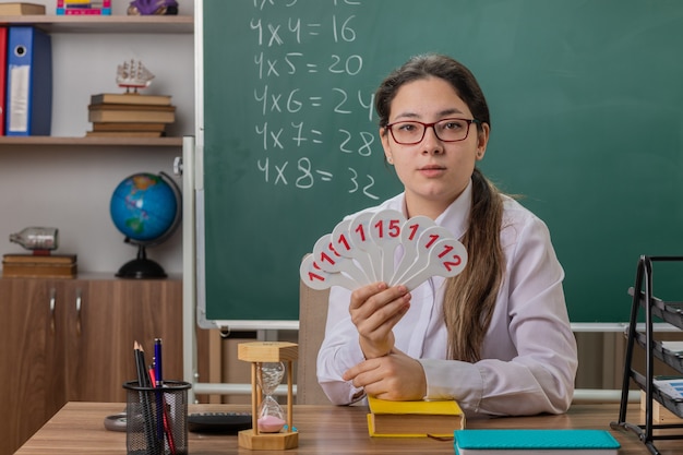 Professora jovem usando óculos segurando placas de matrícula, parecendo confiante se preparando para a aula, sentada na mesa da escola em frente ao quadro-negro na sala de aula