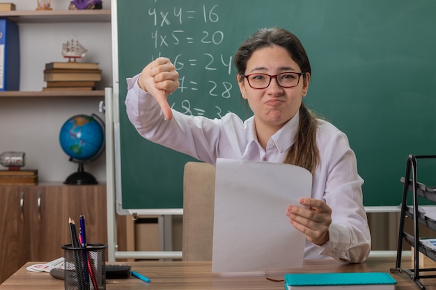 Professora jovem usando óculos, sentada na mesa da escola com páginas em branco, parecendo confusa e descontente mostrando os polegares para baixo, verificando o trabalho de casa na frente do quadro-negro na sala de aula