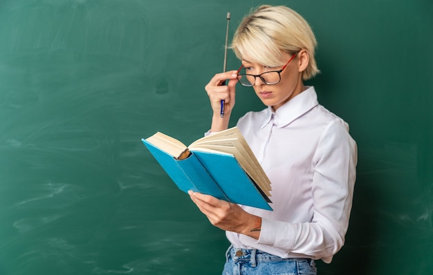 Professora loira jovem concentrada usando óculos na sala de aula em vista de perfil em frente ao quadro-negro, segurando o ponteiro e lendo um livro agarrando os óculos com espaço de cópia
