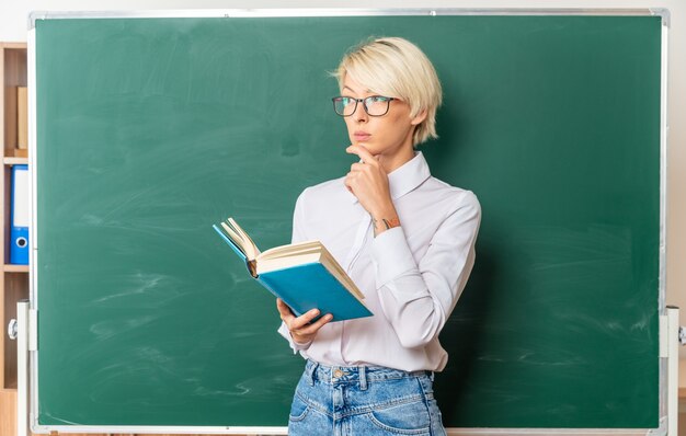Professora loira jovem pensativa usando óculos na sala de aula em frente ao quadro-negro segurando o livro, segurando o queixo, olhando para o lado com espaço de cópia