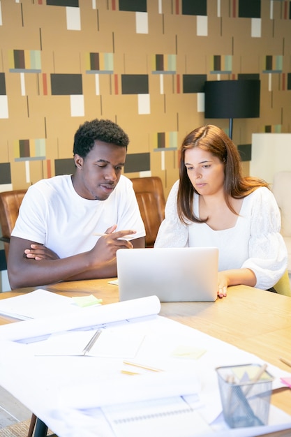 Profissionais criativos trabalhando em projeto de design em laptop juntos, sentados à mesa e conversando