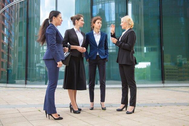 Profissionais femininas de sucesso discutindo o projeto ao ar livre. Mulheres de negócios de terno, juntas na cidade conversando. Comprimento total, ângulo baixo.