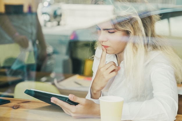 Profissional feminina pensativa sentada à mesa em um espaço de trabalho compartilhado ou café, usando um tablet