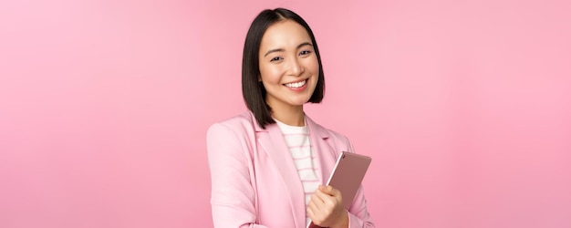 Profissional sorridente empresária asiática em pé com tablet digital vestindo terno para trabalho de escritório parecendo confiante e feliz posando contra fundo rosa