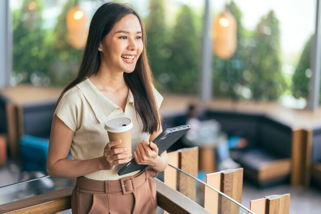 Programador feminino asiático casual codificando estilo de vida digital momad mão segure o tablet e a xícara de café sorrindo alegre sentimento pacífico trabalhando em qualquer lugar em pé no café restaurante reduzindo o estilo de vida