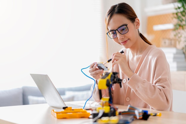 Programadora asiática inteligente aprendendo cabo de placa eletrônica de codificação ai de braço robótico em stem steam, ela tenta testar seu braço robótico autônomo com sensores via plataforma arduino em casa