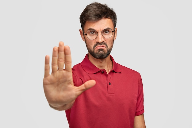 É proibido! Homem jovem irritado e descontente franze a testa, mostra gesto de pare, mantém a palma da mão na frente, tenta se prevenir de algo ruim e desagradável, veste camiseta casual, isolada no branco