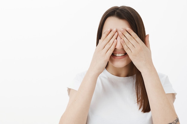 Prometa não espiar. retrato de uma menina animada e feliz aniquilando surpresa com os olhos fechados e as palmas das mãos no rosto sorrindo amplamente como esperando um milagre ou presente, posando alegremente contra uma parede cinza