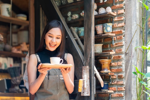 Proprietária do café sorrindo com felicidade positiva pronta para abrir para a mulher feminina customerasian com bandeja de presente de mão de avental e xícara de café condident de boas-vindas na entrada do coffeeshop