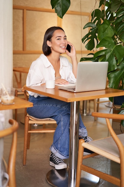 Proprietária jovem gerente de café sentada com laptop atendendo telefonemas e sorrindo amigável