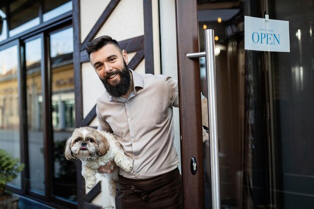 Proprietário de bar feliz segurando um cachorro enquanto abre a porta de entrada e olhando para a câmera