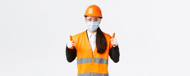 Protocolo de segurança covid19 na construção da empresa e prevenção do conceito de vírus engenheira industrial asiática confiante em máscara facial e capacete mostrando o polegar para cima tudo de bom