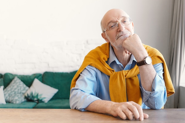 Psicólogo sênior careca com óculos retangulares e suéter amarrado nos ombros, sentado na mesa de madeira vazia do escritório em casa, esperando pelo cliente, com expressão facial atenciosa