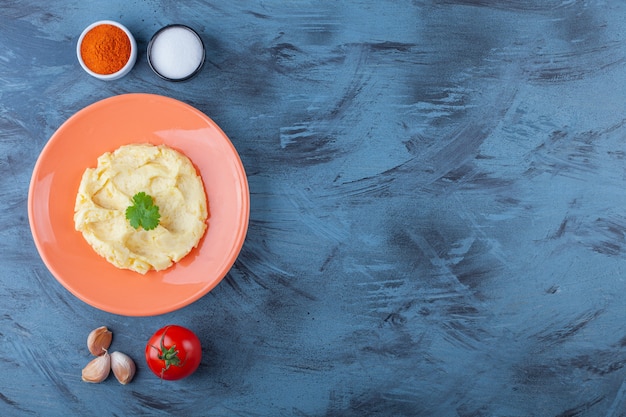 Purê de batata em um prato próximo a vegetais e tigelas de temperos na superfície azul