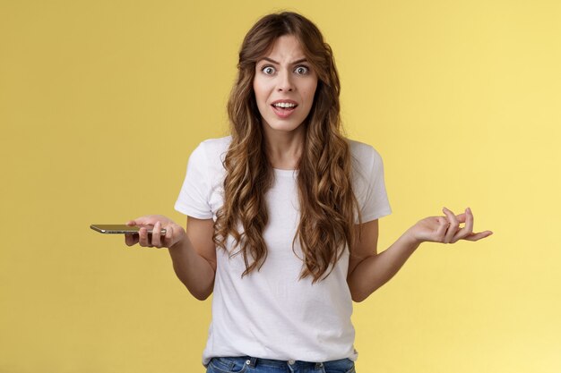 Puto frustrado incomodado menina caucasiana de cabelos cacheados olhar câmera perplexo reclamar segurar smartphone encolher os ombros espalhar as mãos para o lado desânimo discutindo após intenso fundo amarelo de telefonema sério.