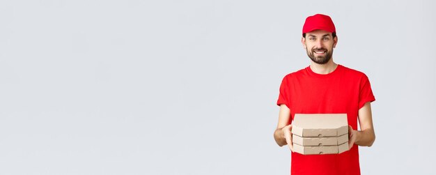 Foto grátis quarentena de entrega de alimentos fique em casa e peça o conceito on-line correio barbudo sorridente com boné uniforme vermelho e camiseta entregando caixas aos clientes com fundo cinza pedido de entrega de pizza