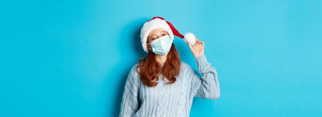 Foto grátis quarentena de natal e conceito cobiçoso alegre adolescente ruiva com chapéu de papai noel e máscara facial encarando