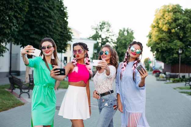 Quatro melhores amigas elegantes e lindas de óculos, cada uma posando fazendo selfies para as redes sociais, se divertem. Mulheres em óculos de sol depois de fazer compras e salão de beleza.