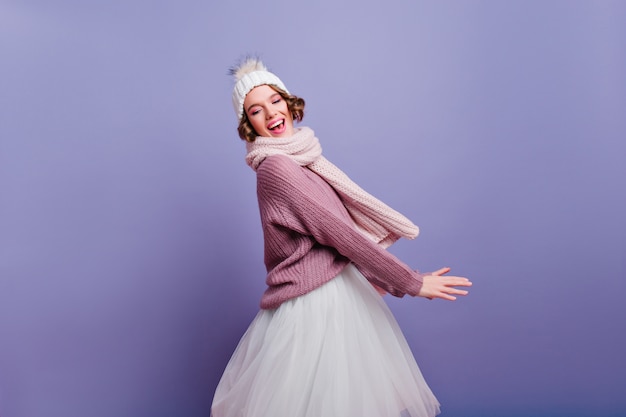 Que bom que garota com cabelo curto e brilhante, posando com um lindo chapéu branco. dançando atraente modelo feminino com saia e lenço de malha.