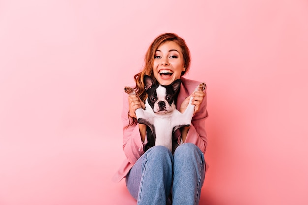 Que bom que garota de jeans brincando com um cachorrinho engraçado. Retrato interno da senhora ruiva animada com penteado encaracolado, passando um tempo com seu cachorro.