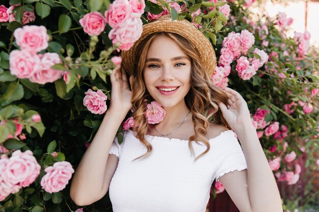 Que bom que garota de olhos azuis posando na natureza e sorrindo. Impressionante modelo feminino no moderno chapéu de palha, relaxante sabor de flores.