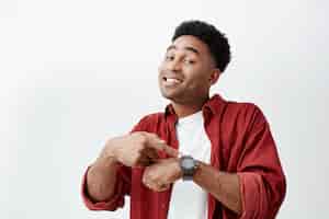 Foto grátis que horas são. retrato de jovem homem atraente de pele escura com penteado afro escuro em camiseta branca e camisa vermelha, apontando o relógio à mão com a expressão do rosto feliz, mostrando a hora de comer.