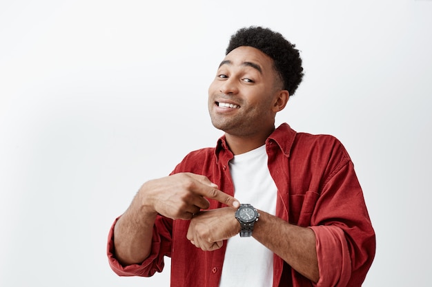 Que horas são. Retrato de jovem homem atraente de pele escura com penteado afro escuro em camiseta branca e camisa vermelha, apontando o relógio à mão com a expressão do rosto feliz, mostrando a hora de comer.