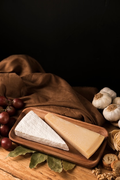 Queijo, alho, uvas e folhas de louro na bancada de madeira contra o pano de fundo preto