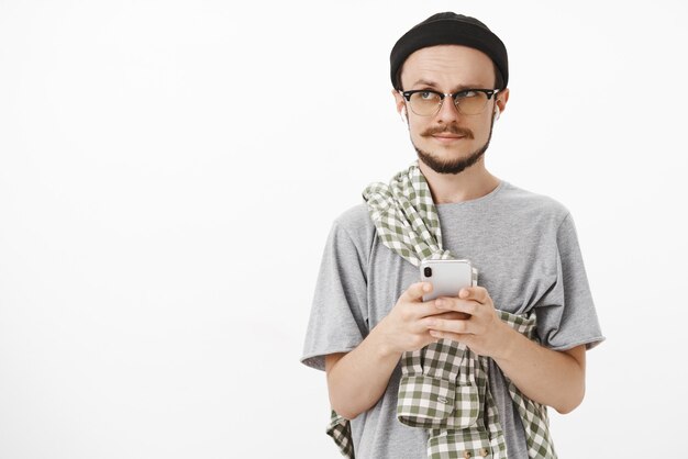 questionou artístico bonito barbudo homem com gorro preto e camisa cruzada no peito segurando um smartphone levantando uma sobrancelha enquanto olha para a esquerda com uma música confusa em fones de ouvido sem fio