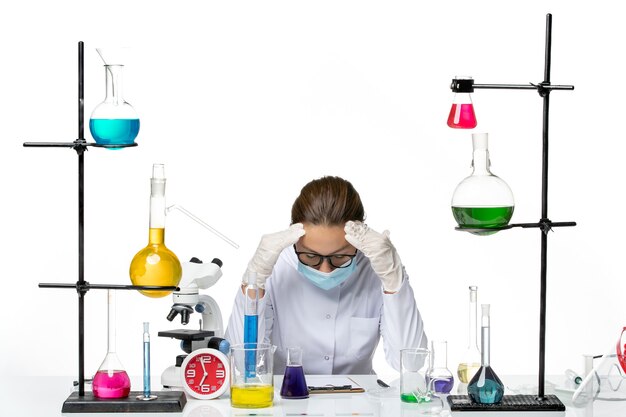 Química feminina vista frontal em traje médico com máscara sentada em frente à mesa com soluções cansadas no fundo branco vírus químico laboratório respingo secreto