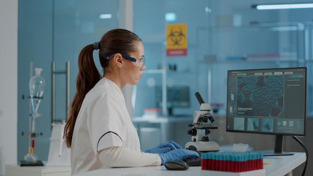 Químico com óculos de proteção e luvas trabalhando no computador para analisar animação de dna para desenvolvimento de biologia. mulher olhando para o monitor com ilustração de diagnóstico científico.