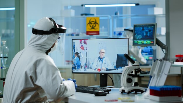 Químico em terno de ppe ouvindo médico profissional em videochamada, discutindo durante reunião virtual no laboratório de pesquisa. Médicos usando alta tecnologia para pesquisar tratamento contra o vírus covid19