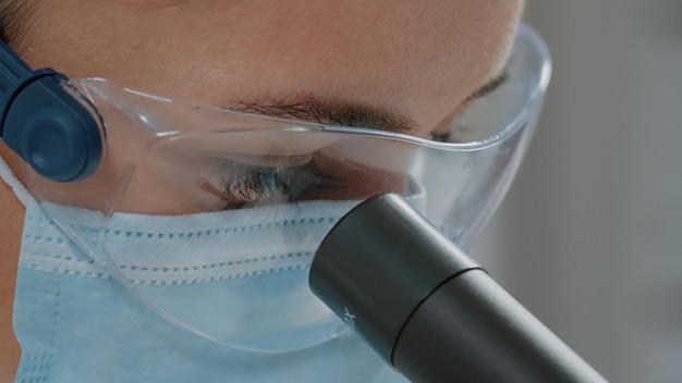 Químico olhando através de lentes microscópicas na ferramenta óptica, usando máscara facial e óculos de proteção em laboratório. microbiologista usando microscópio e foco seletivo para experimentos. fechar-se
