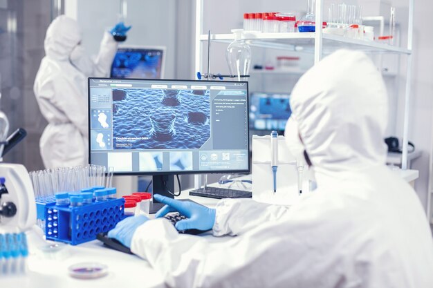 Químico sentado em seu local de trabalho em laboratório analisa coronavírus no computador vestido de ppe