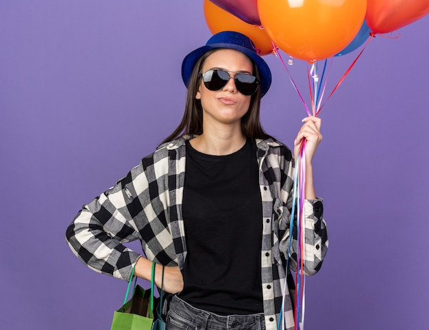 Rapariga bonita com chapéu de festa e óculos segurando balões e colocando a mão no quadril