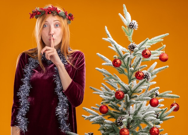 Rapariga bonita em pé perto da árvore de Natal com vestido vermelho e grinalda com guirlanda no pescoço, mostrando gesto de silêncio isolado em fundo laranja