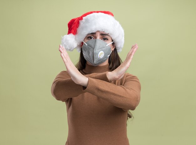 Foto grátis rapariga bonita usando chapéu de natal com máscara médica mostrando gesto de não isolado em fundo verde oliva