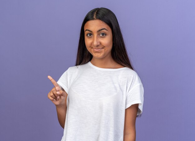 Rapariga com uma t-shirt branca a olhar para a câmara a sorrir e a apontar com o dedo indicador para o lado, de pé sobre um fundo azul