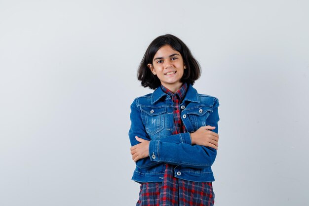 Rapariga em pé de braços cruzados em camisa xadrez e jaqueta jeans e olhando bonito, vista frontal.