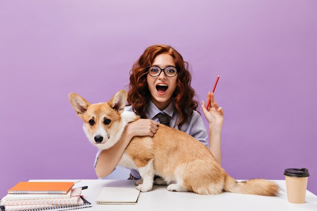 Rapariga encaracolada com camisa e óculos a abraçar cão e a segurar um lápis