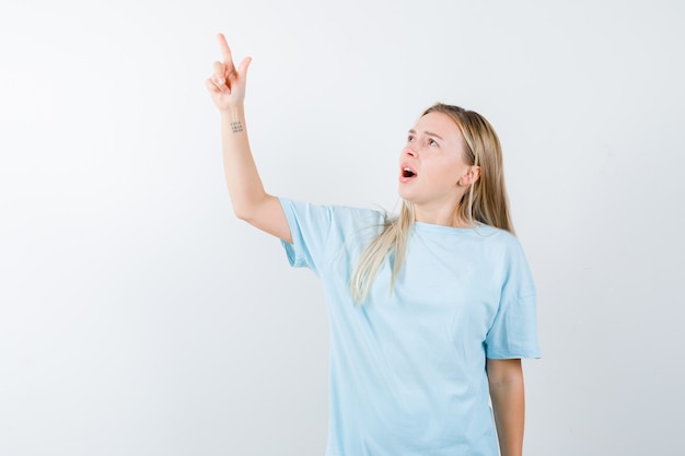 Rapariga loira com t-shirt azul apontando para cima com o dedo indicador e olhando surpresa, vista frontal.