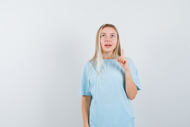 Rapariga loira com t-shirt azul apontando para cima com o dedo indicador, olhando para cima e olhando focada, vista frontal.