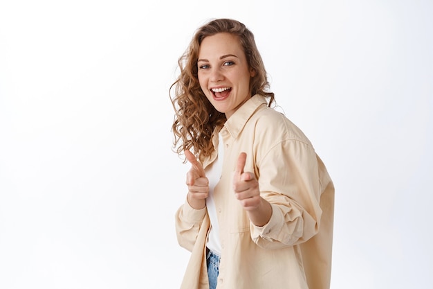 Rapariga loira sorridente anima, elogiando ou escolhendo você, apontando o dedo para a frente e parecendo feliz em pé sobre uma parede branca