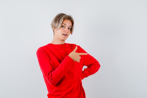 Rapaz adolescente apontando para o lado direito com um suéter vermelho e parecendo surpreso. vista frontal.