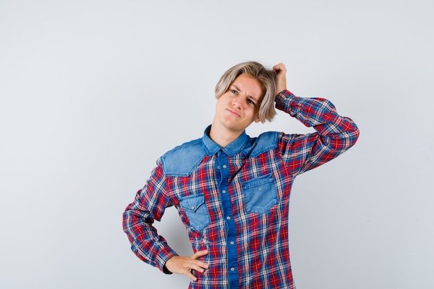 Rapaz adolescente coçando a cabeça enquanto olha para o lado em uma camisa quadriculada e parece pensativo. vista frontal.