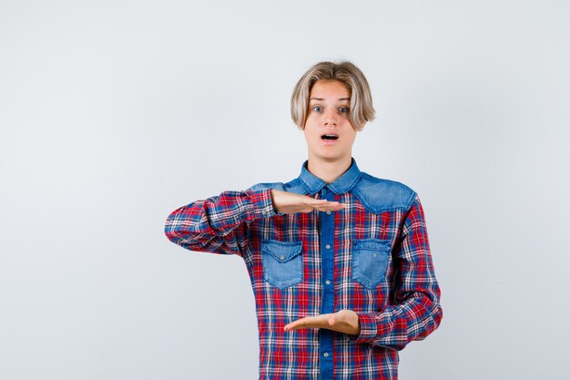 Rapaz adolescente com camisa quadriculada, mostrando sinal de tamanho e olhando perplexo, vista frontal.