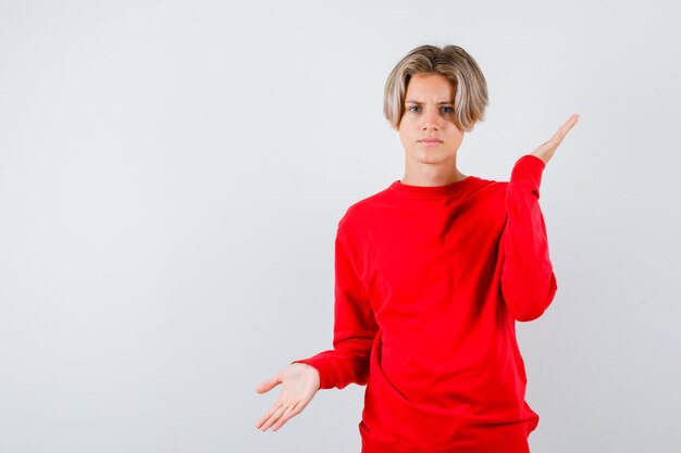 Rapaz adolescente com suéter vermelho, mostrando um gesto impotente e olhando indeciso, vista frontal.