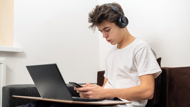 Rapaz adolescente está usando smartphone com fones de ouvido, laptop de joelhos em casa. Rosto sério e focado